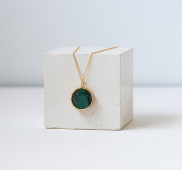 Round emerald pendant on a gold chain draped over the white cube.