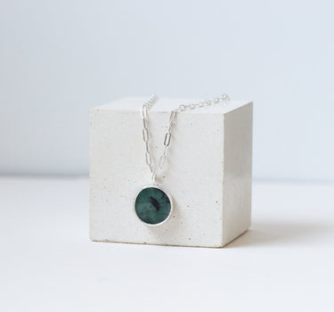 Round emerald pendant on a silver chain draped over the white cube.